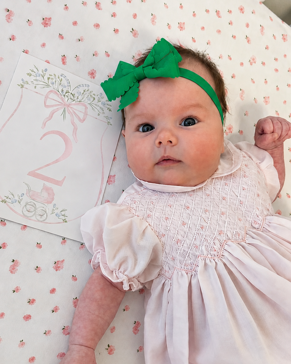 A baby lying on a patterned blanket wearing a white smocked dress, a bright green bow headband, and looking at the camera. A pink and white card is tucked under the baby's head.