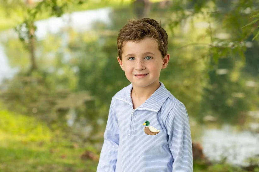 A young boy with brown hair stands outdoors in front of a blurred lake and trees, wearing a light blue and white striped quarter-zip polo shirt with a small embroidered mallard duck logo on the left chest.