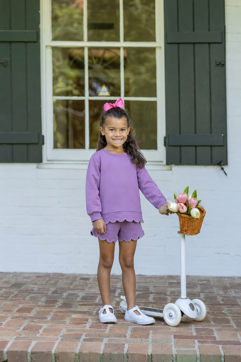 A young girl stands on a brick porch in front of a white wall with dark green shutters, wearing a matching lavender-purple long-sleeve top and shorts set with scalloped edges. She holds a small basket filled with flowers on a white scooter.