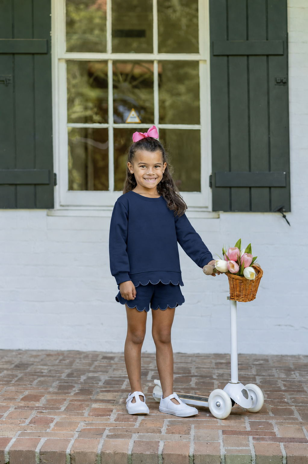 A young girl stands on a brick patio in front of a white house with dark green shutters, wearing a navy blue long-sleeve top and matching shorts with a pink bow in her hair. She is holding a small basket of flowers next to a white scooter.