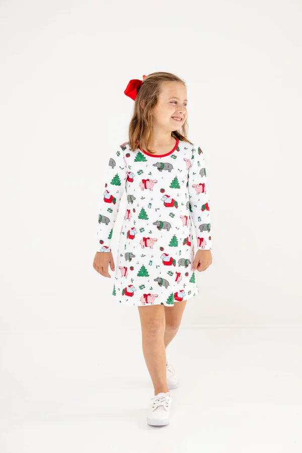 A young girl wearing a white long-sleeve holiday-themed dress with a print of Christmas trees, Santa Claus, and snowman, standing against a plain white studio background.